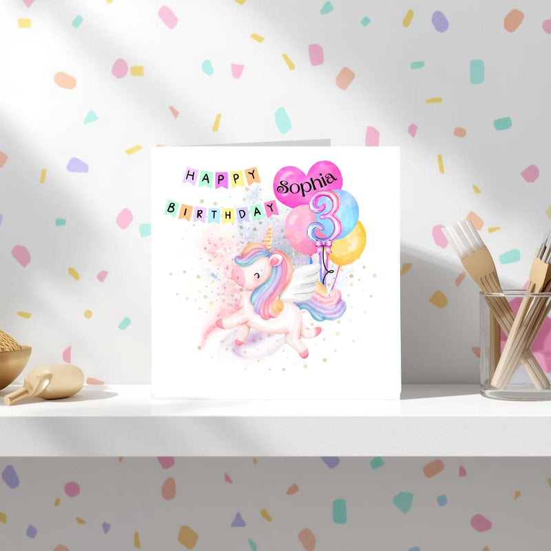 May include: A white birthday card with a pastel unicorn illustration. The card features a cluster of balloons with the number 3 and the name "Sophia". The words "Happy Birthday" are written above the unicorn in colourful letters.