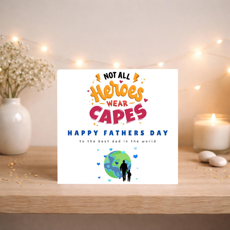 May include: A white Father's Day card with the text "Not All Heroes Wear Capes." The card features a globe with a silhouette of a father and child. The card is on a wooden surface with a vase of flowers and a lit candle.